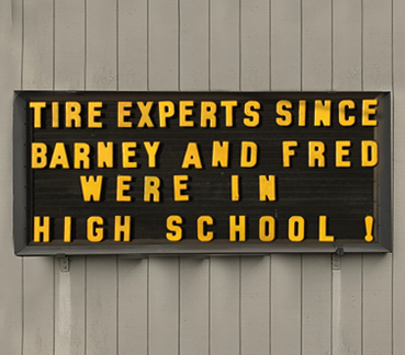 Outdoor sign with text: TIRE EXPERTS SINCE BARNEY AND FRED WERE IN HIGH SCHOOL!